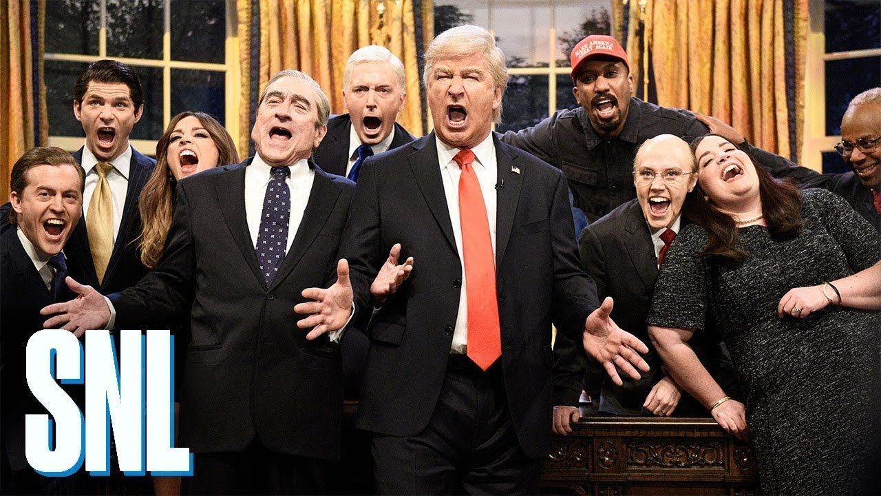 An scene featuring the cast of Saturday Night Live in the Oval Office, with Robert Deniro and Alec Baldwin as Donald Trump in the foreground.