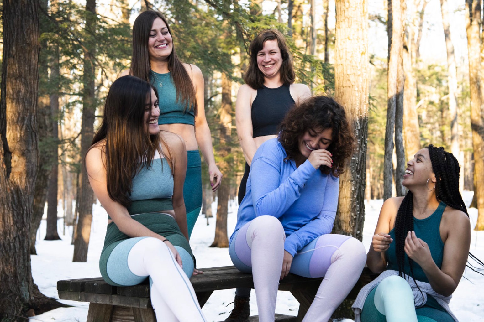 Five women wearing athletic wear laughing on a picnic table in the woods. Snow is on the ground.